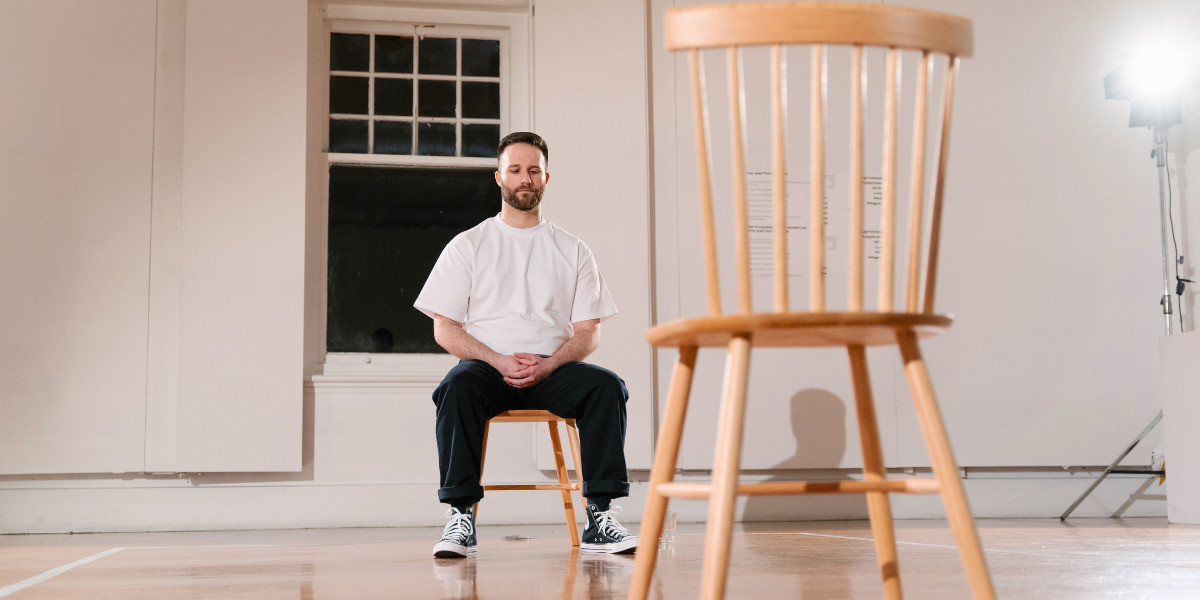 A man in a white t-shirt, black pants, and black and white Converse shoes sits calmly on a wooden chair opposite an empty wooden chair.
