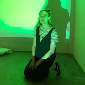CANCELLED - Woolgatherer - Steph kneels on a concrete floor wearing blue overalls and a striped shirt. She is sitting in front of a projection that turns herself and the blank wall behind her green.