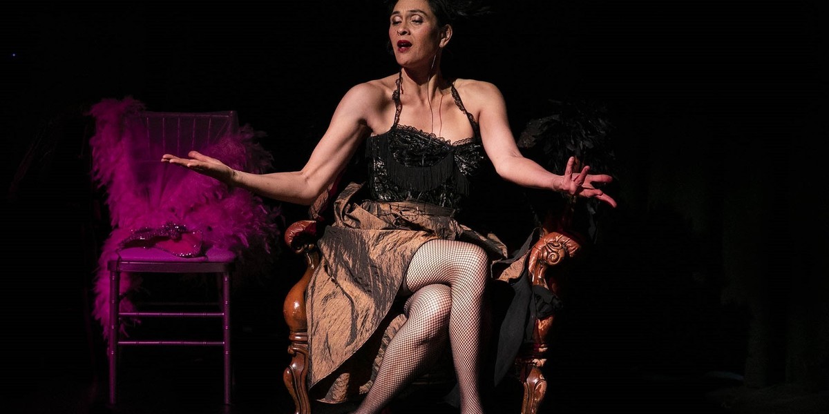 Woman in black feathered headdress sits on stage in a comfortable old armchair, legs crossed, arms out, animatedly telling a story. Behind her is a pink feather boa on a chair. She is wearing a corset, rose gold shoes, fishnet tights and a bustle skirt. Photograph by Andrew Worssam.