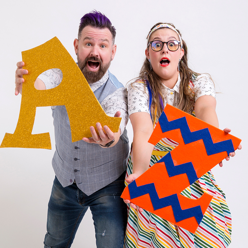 A man with purple streaked hair holds a large sparkly gold coloured "Letter A" next to a woman in a striped dress holding a large orange and blue "Letter Z".