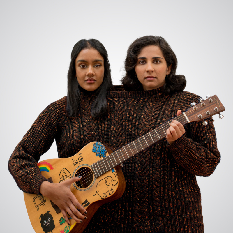 Leela and Shabana are standing in front of a white background. Both are wearing the same oversized jumper with their heads coming out of the same hole. They have very blank serious facial expressions. Shabanas' hair is black, parted in the middle and straight. Leelas' hair is parted to the side and wavy/curly. Shabana holds the base of a guitar and Leela holds the neck. The guitar is acoustic, wooden and covered in drawings and stickers of a rainbow, an old fashioned tv, an elephant, eyes and a little monster.