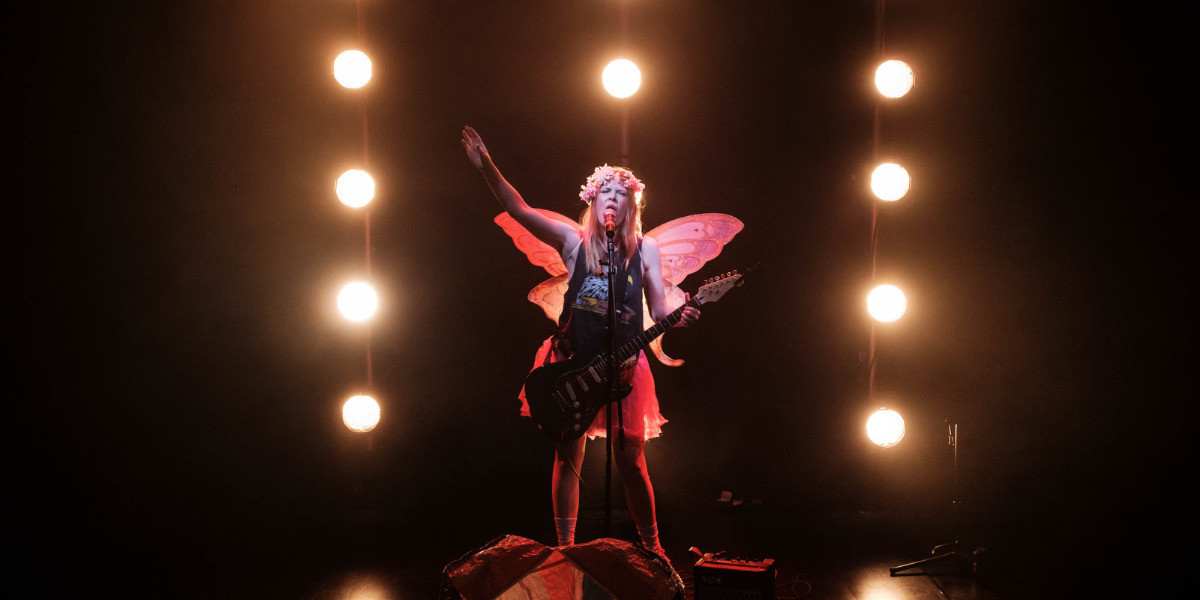 a woman wearing party wings stands in front of a wall of rock lights playing guitar