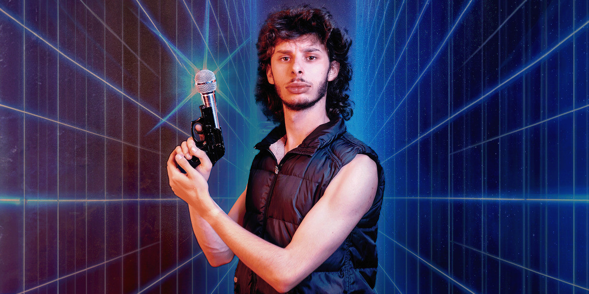 CON 2077 - A young mulleted man wearing a black singlet holds a microphone that looks like a gun. He stands in front of a neon grid background.