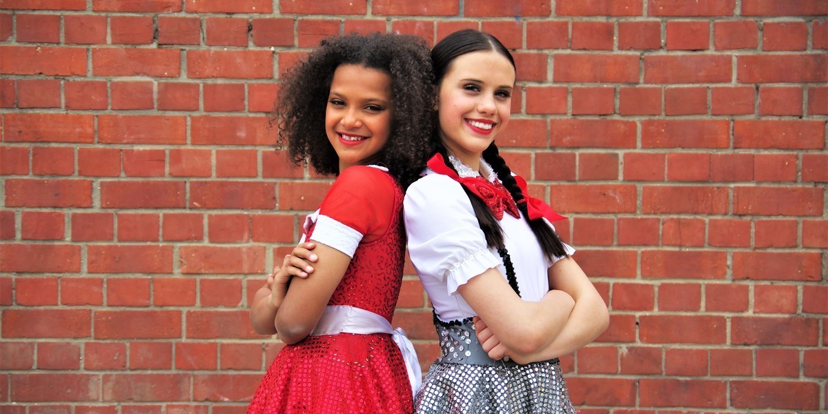 Annie in a bright red dress stands back to back with Matilda in a school uniform against a red brick wall.