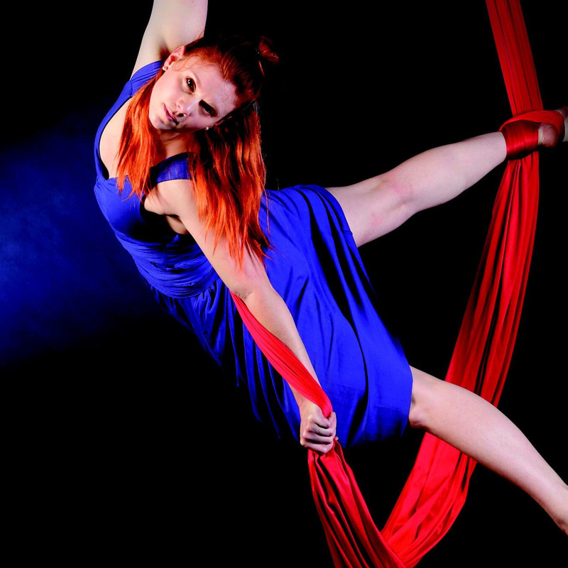 Aerial Silks Workshops for Adults - An aerial artist in a purple dress with long orange hair and fair skin is suspended between two red ribbons. Her right foot is intertwined with the ribbon while her left one hangs down, Her right arm holds the top of other ribbon and her left arm grasps the bottom end of the ribbon, suspending her body sideways. Her head is lifted facing the front. The background is dark grey.