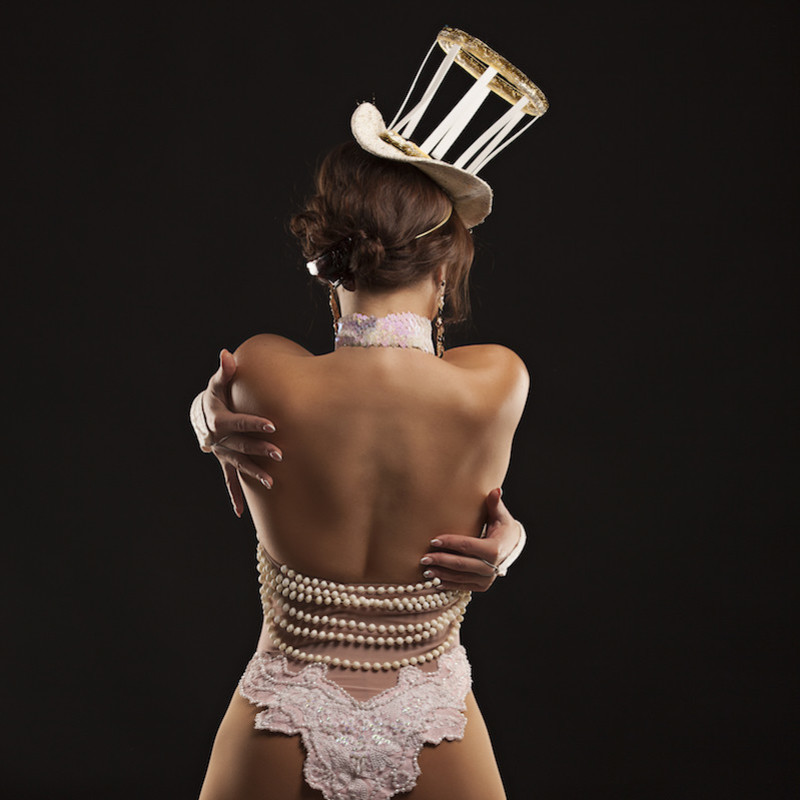 Woman with her back to the camera wrapping her arms around herself, in a top hat and matching lingerie