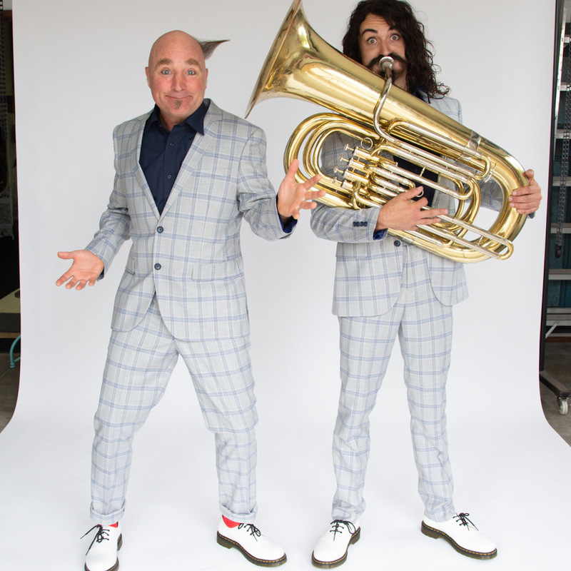 Two men are looking directly at the camera, one man is smiling the other is blowing a trombone with wide eyes.