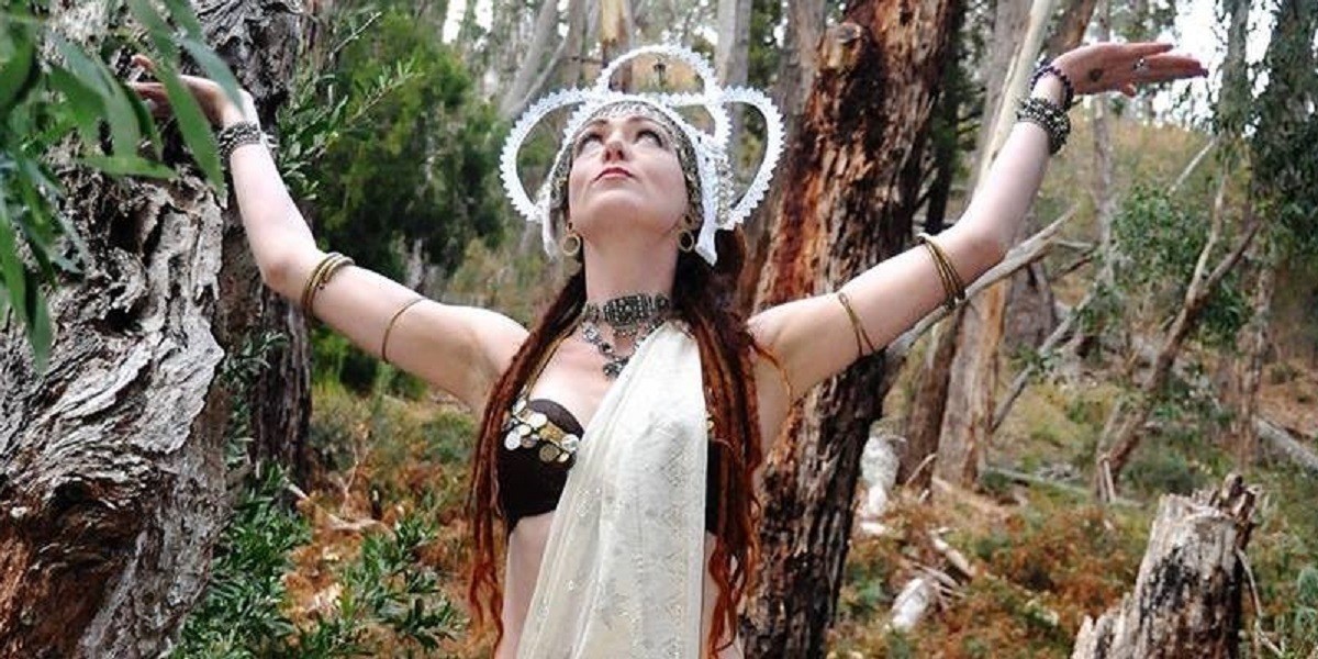 A photo of a woman standing amongst a forest. She is wearing a black top and a white sash across her body. Her arms are in the air and she is wearing several bangles along her arms. On her head she is wearing a white decorative headpiece.