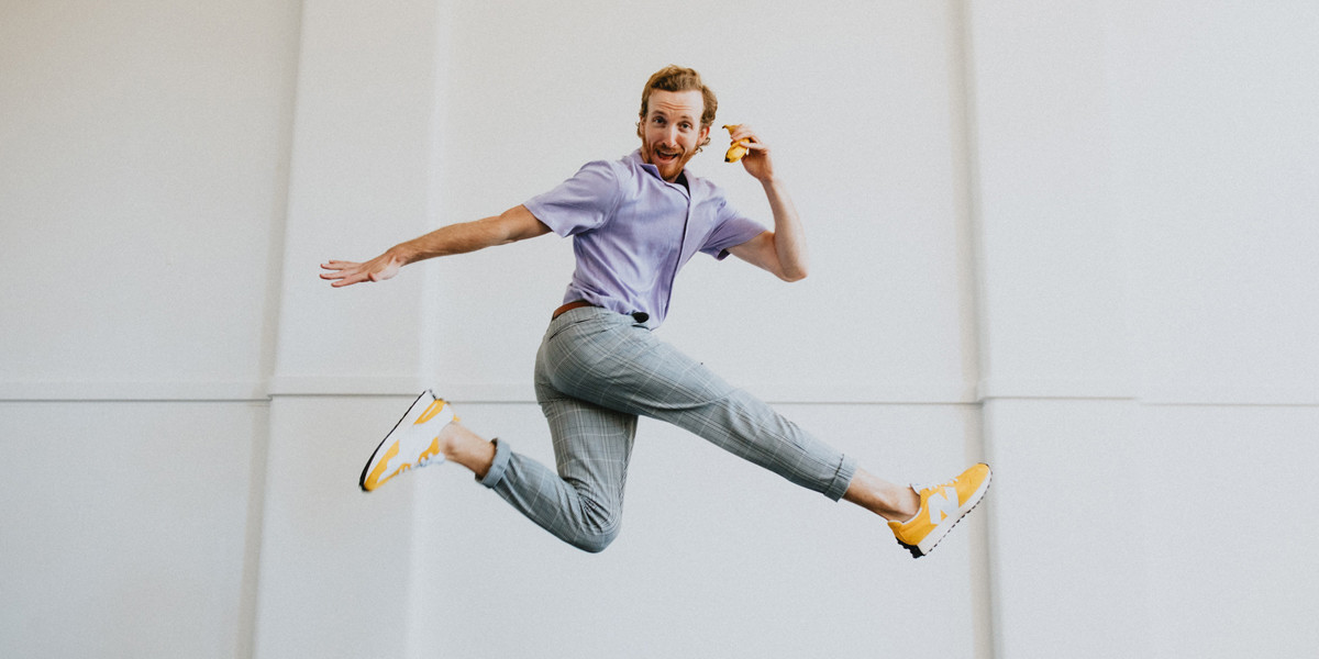 Will jumps high in the air, with a joyous expression. In one hand he holds a banana as if it's a phone, the other outstretched behind him. He is wearing a pastel purple polo shirt, bright yellow shoes, and grey striped suit pants, against a white wall.