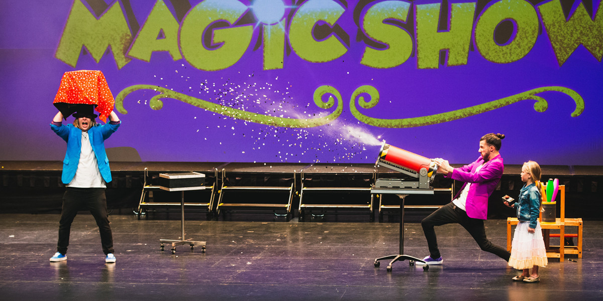 two male performers, one firing a red cannon shooting confetti across a stage.