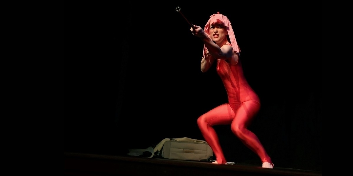 Sarah-Louise Young is dressed in a red lycra jumpsuit. Half squatting, she's holding a mop with both hands pointing it towards someone in the audience (out of shot), she wears the pink mop-head as a wig.