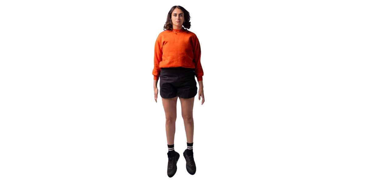 Out of Body Experience - A plain white background with a young woman floating upright in the centre. Her feet are floating in the air and her hair bounces around her face thanks to gravity. She has tanned Caucasian skin, brown curled hair that goes to shoulders, and is wearing an orange crewneck jumper, a plain black skirt, black socks and black running shoes. Her facial expression is blank and she looks directly into the camera with a neutral stare.