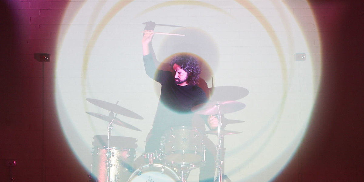 Drummer plays behind graphics projected onto a transparent screen in front of the drum kit