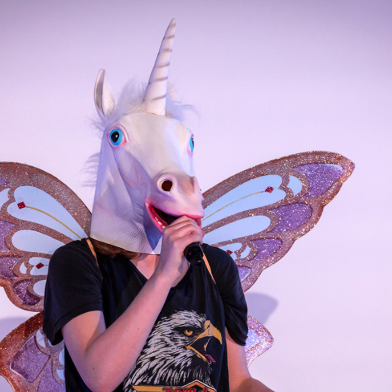 a woman in fairy wings, a black t-shirt and a unicorn mask holds a mic to the masks mouth.