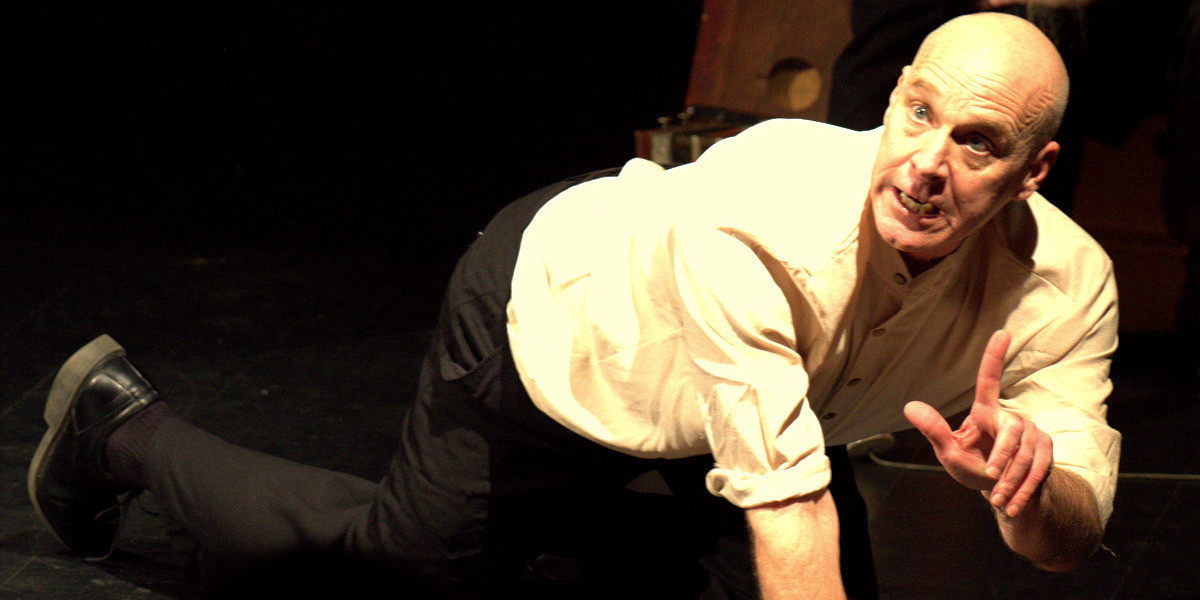 Under stage lights, a man crawls on the floor while with one hand, he lifts a single finger as if pausing to make some important point.