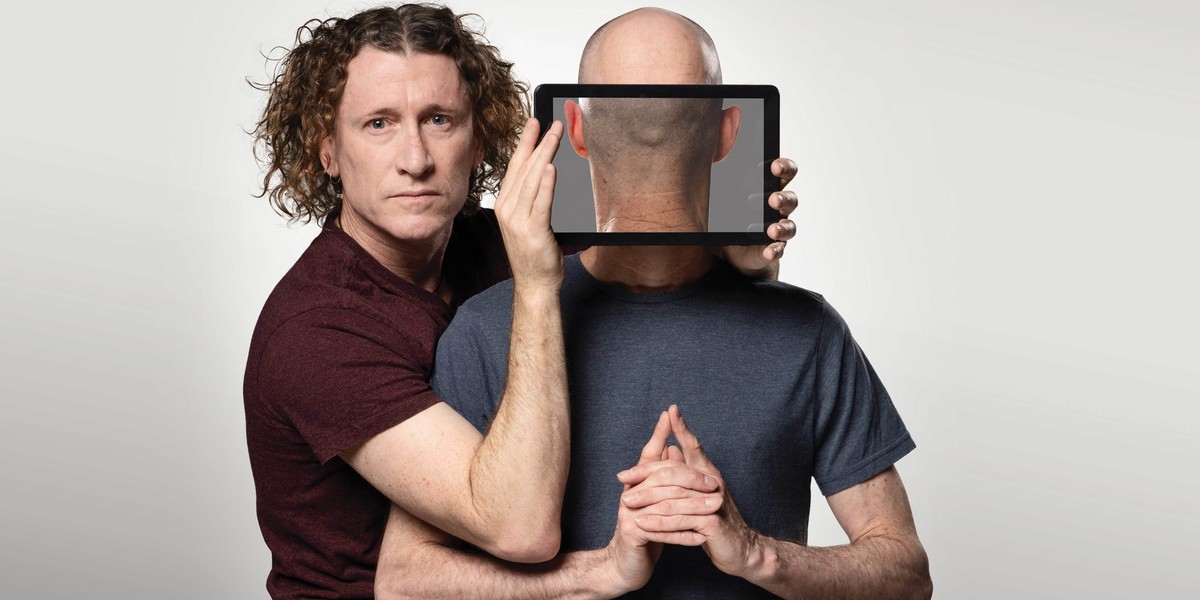 The Umbilical Brothers - David on the left is in a maroon shirt, facing the camera holding an ipad which shows the back of the man on the right's head (Shane) although his body is facing forward.