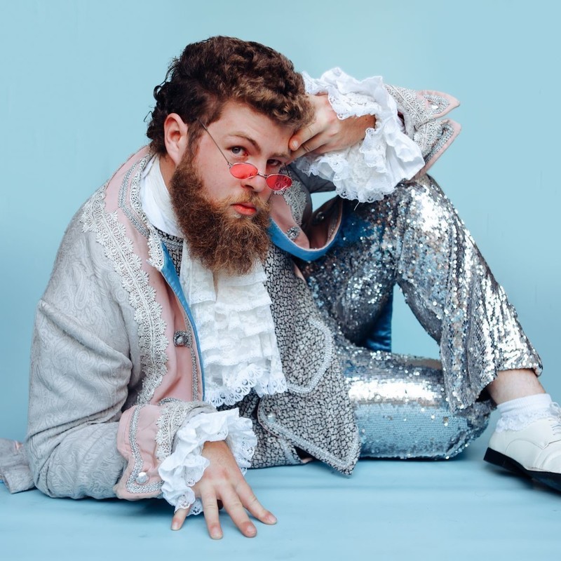 A man wearing a baroque jacket and sparkly disco flare pants poses strikingly on the ground.