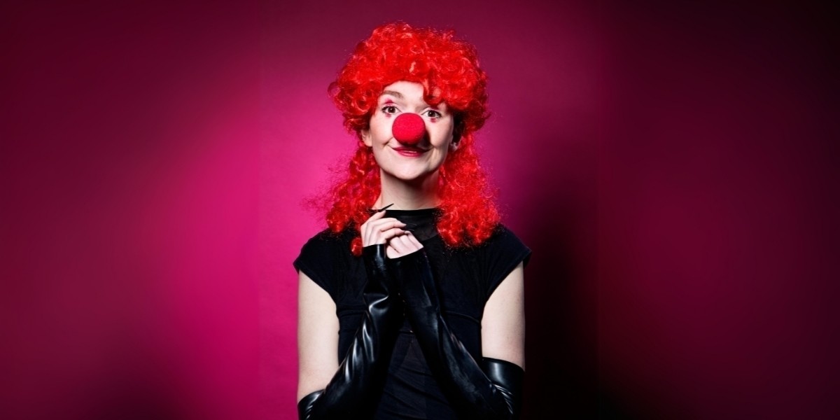 Elf - dressed in a black cap-sleeved dress and black fingerless gloves - looks directly at the viewer, shoulders squared to the camera. Her hands are clasped innnocently in front of her collarbones, and her face - adorned with traditional white-faced clown make-up (including a foam red nose) - wears an angelic expression. She's wearing a big bright red, curly shoulder-length wig.