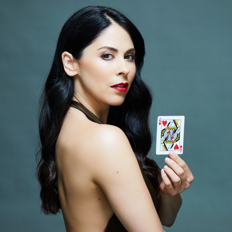 The female magician hold a playing card in her hand. She has long dark brown hair and a green dress with red lipstick on her lips.