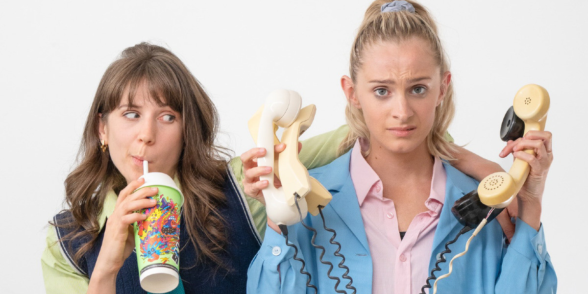 A brunette girl drinks a slurpee and looks quizzically at a blonde girl, holding many old phones up to her ears with an expression of concern.