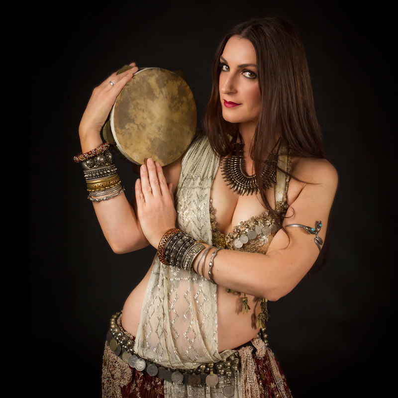 A woman wearing a beige belly dancing costume with silver embellishments. She has a cream coloured sash across her body. She has multiple bangles around each wrist and is holding small hand drum over her shoulder.