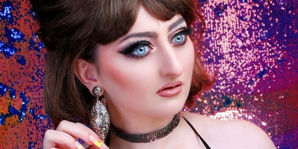 I'm Not A Robot - Close up of comedian, Sonia Di Iorio wearing a wig, glo mesh top, choker and glitter earrings. She has heavy eye make-up and looks dazed. She is standing in front of a glitter backdrop.