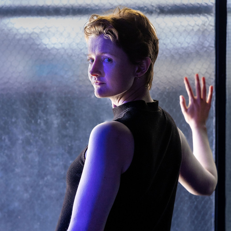 Lucy, a white person with short light brown hair and blue eyes, looks back over their left shoulder at the camera. She is standing in front of a window with her right hand flattened against the glass. Only gray light and indistinct shapes can be seen through the frosted glass.