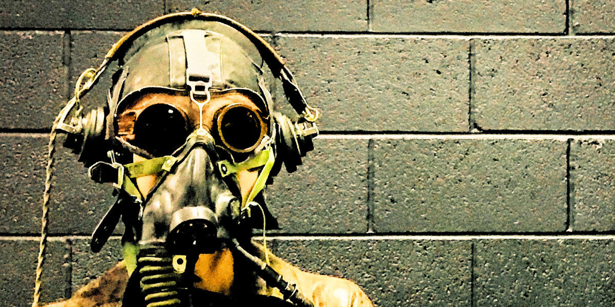 Bunker - A person in a protective suit and steampunkish gas mask leans against a grey brick wall in a bunker.