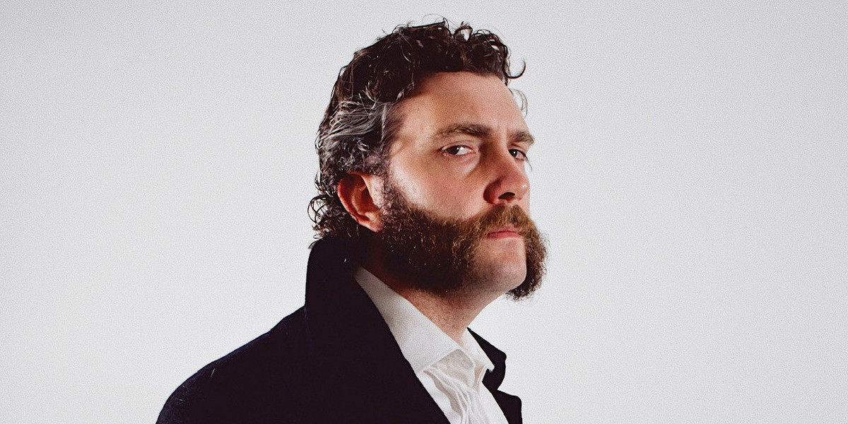 A 3-quarter side profile of Jacob Jackman against a white background. He has a shaggy, greying mullet and robust mutton chops. There are bags under his eyes and a serious expression on his face. He is wearing a white tuxedo shirt and thick black overcoat.