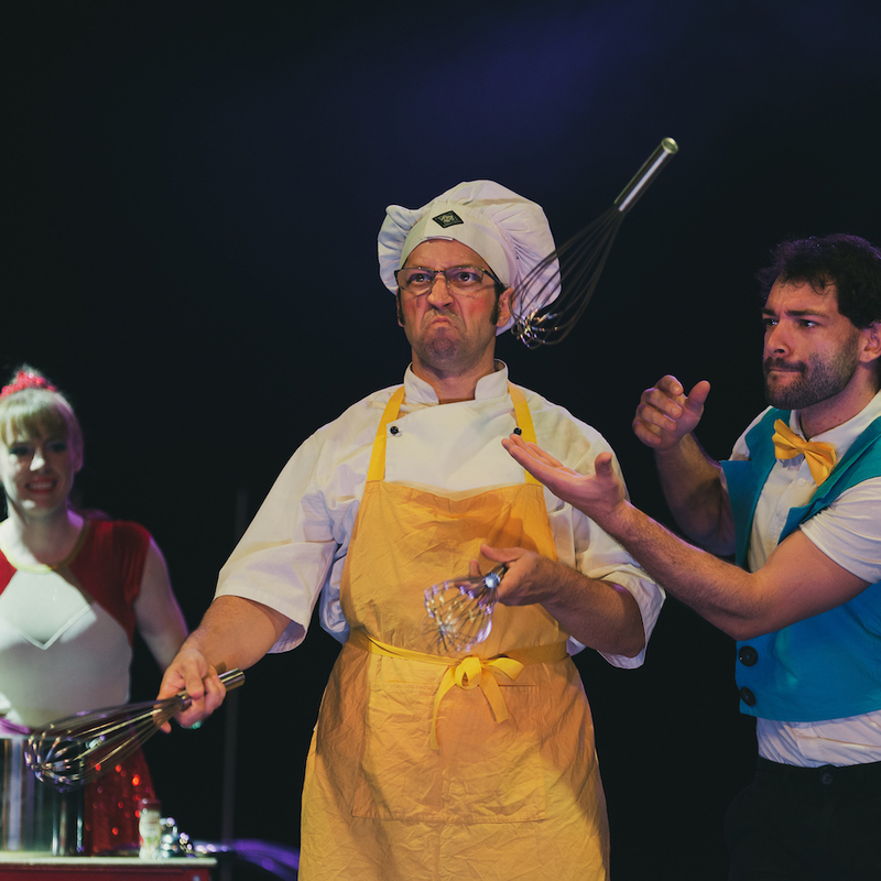In the centre is a man in a chef hat and yellow apron frowning while juggling kitchen whisks as one is stolen by a man on the right in a blue tails jacket. A woman in a red and white shirt stands behind them watching.