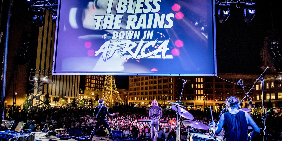 A large screen takes up the upper mid-ground emblazoned with the text "I bless the rains down in Africa". The camera angle appears to be positioned at the back of a deep stage, in the foreground to the right we see the back of a drummer wearing a black tank top, hair in a messy brunette ponytail, mid-play. To the drummer's left further down-stage we see the back of a keyboard player dressed in a textured t-shirt and light coloured fitting pants, he is playing a pedal with his left foot. To the right of him is a guitar player, legs akimbo dressed in a shiny jacket and pants. Some other speakers and roadcases appear in the bottom left corner of the stage. The stage faces out on a large crowd of people, some in the centre are well lit, others are shadows as it is nighttime. They are surrounded by a high-rise building on the left and lower multi story buildings in the centre and left hand side.