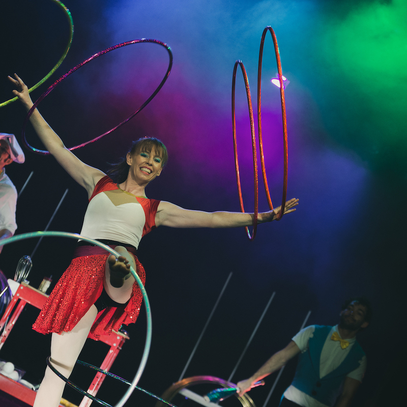 A woman wearing a red skirt looks at the camera smiling while she stands on one leg with six hula hoops spinning on her body. In the background a man wearing a yellow apron and chef hat is walking.