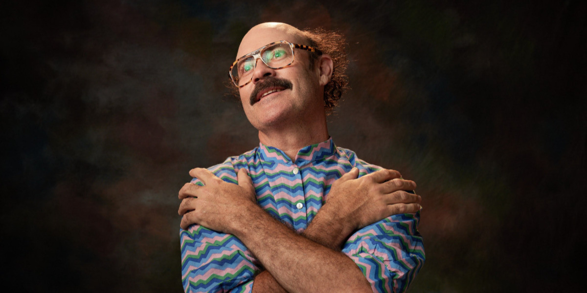 Sam Simmons - Man With A Fork In A World Full Of Soup - Sam is wearing a zig zag pattern button up shirt. The shirt is pink, blue and green. He is also wearing brown tortoise shell glasses. He has a brown moustache and brown hair. He is crossing his arms to the opposite shoulder and looking up out camera.