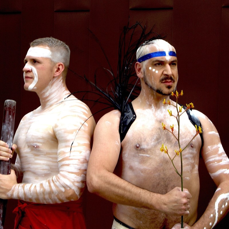 Two First Nation Aboriginal male performers standing back to back both looking away into the distance. Left male has white ochre painted traditionally on body and face and is holding a didgeridoo. Right male artist is painted in white patterns with a blue glittered headband around the head and is holding in both hands a red/ yellow kangaroo paw native flower bunch