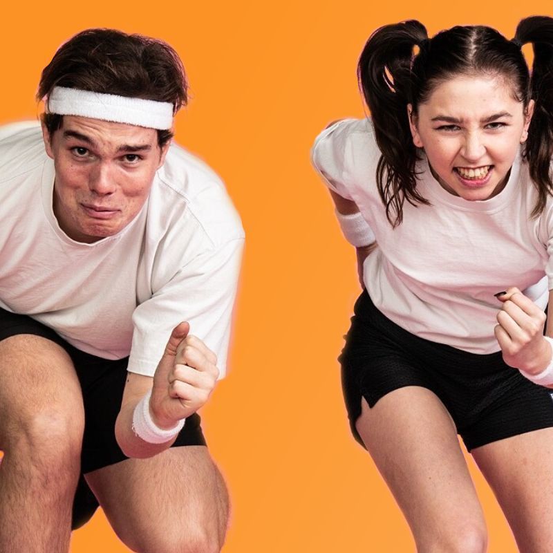 The Beep Test - Two people dressed in black running shorts and white t-shirts hunched over preparing to run in front of an orange background. The male on the left is a caucasian young adult with an incredibly worried expression on his face, he is also wearing a white sweatband on his head and wrists. The female on the left is a caucasian young adult with a determined and angry expression, her hair is in pigtails and she is wearing white sweatbands on her wrists.