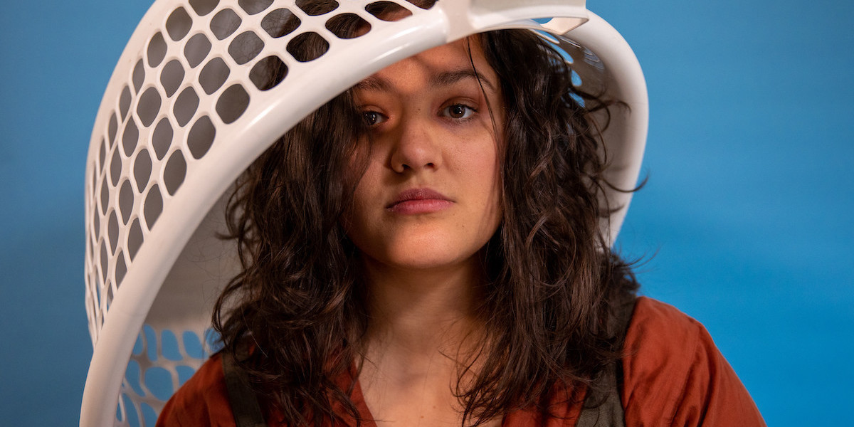 A photo of a woman with brown wavy hair and a blank expression on her face with a white laundry basket on her head.
