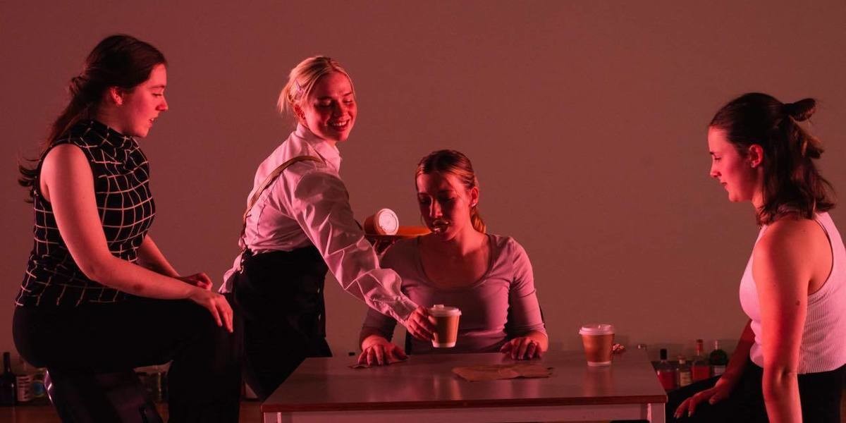 3 people sit around a table in a light pink light setting as a waitress serve them coffee
