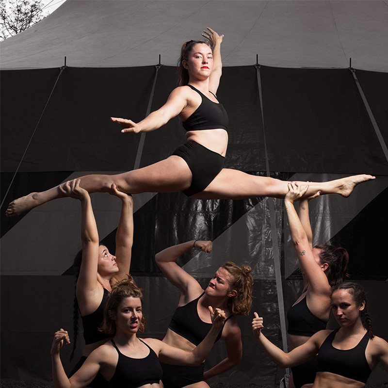 The all-female cast of YUCK Circus performing an acrobatic skill. A performer is lifted in the air in a front spilt, and faces the camera. Below her, the rest of the cast pose with their hand is a shukka position. The image is dark, and against the backdrop of a circus tent