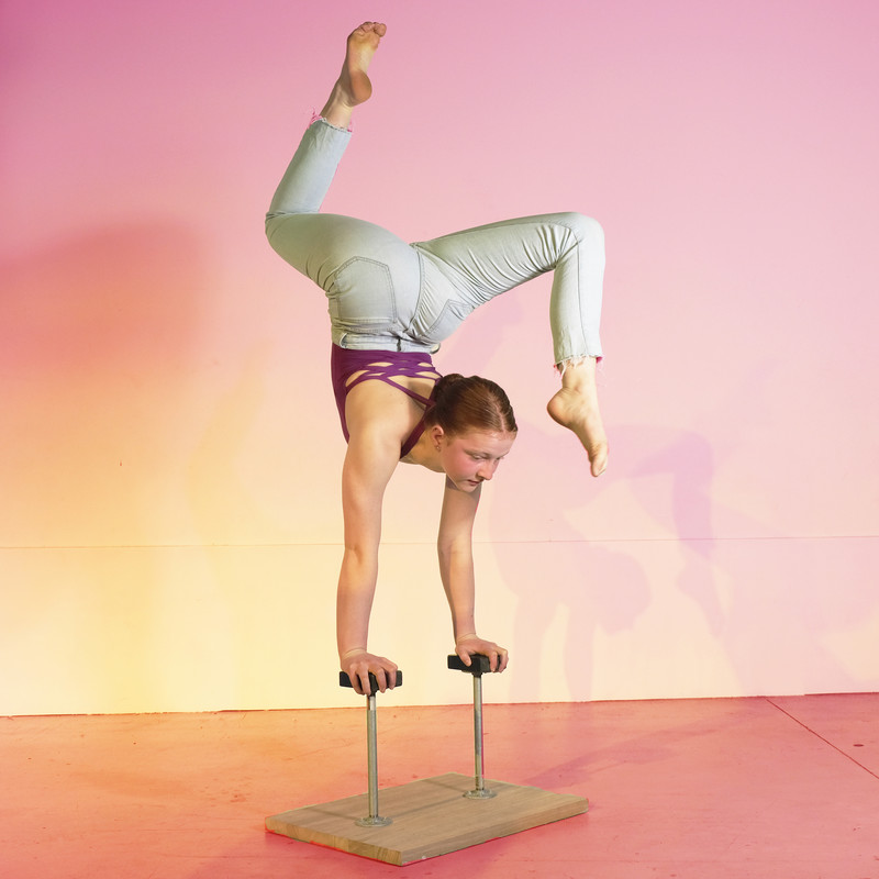 Young Woman does handstand on canes in a double stag position with warm yellows and pinks in the background.