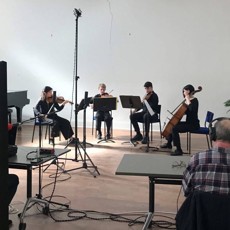 Four musicians play together as a string quartet in a light room with recording equipment surrounding them.