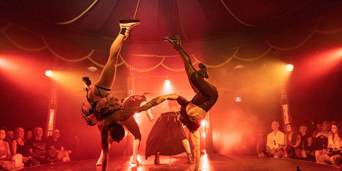 Two people do a breakdance style handstand while holding hands, with an array of performers behind them in a red light.