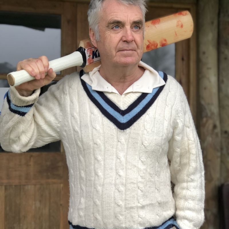 Man standing in white cricket uniform with cricket bat over his shoulder, ready to go out to play.