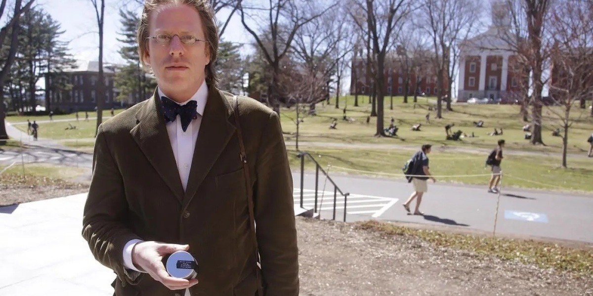 Bestiaries of the More-Than-Humane - A nerdy man stands on a US college campus, blonde hair and glasses, wearing a tweed suit, holding an unspecified device and looking at the camera