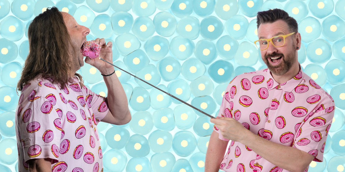 One man with long hair is wearing a pink shirt and standing in profile while eating a doughnut, while another smiling man wearing yellow framed glasses and a pink shirt points at the doughnut with a silver pointer.