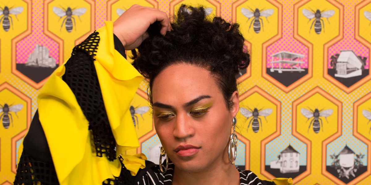 A close-up photographic portrait of a person looking towards the ground, with one hand gesturing towards their hair, in front of a yellow and black graphic background featuring bees and architectural forms in a honeycomb pattern.