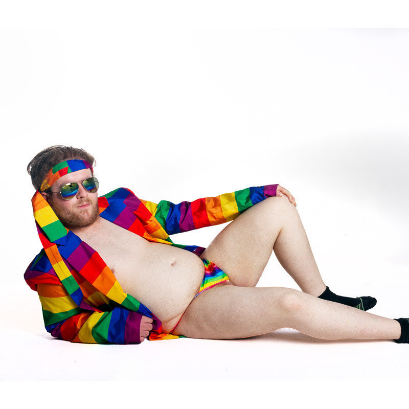A semi naked man reclines on the ground.  He is wearing a rainbow coloured jacket and a rainbow G -String.