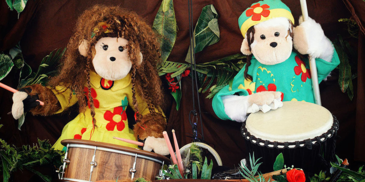Amazing Drumming Monkeys - 20 Year Anniversary Show - Puppet monkeys paying drums.  Girl monkey in yellow, on left of picture, playing snare drum with sticks.  Boy monkey in green on right of picture, playing African djembe drum with hands.
