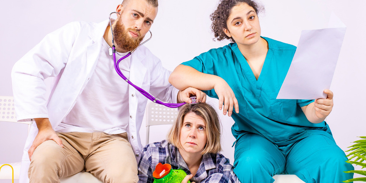 Three white young adults sit in a staged medical waiting room. To the left, Maxx who wears a medical coat holds a purple stethoscope to Abbie’s head, who is sat on the ground holding a hot water bottle. To the right, Emily with dark curly hair sits on a chair reading medical notes and leans her arm on Maxx’s stethoscope, looking into the camera with a blank expression.