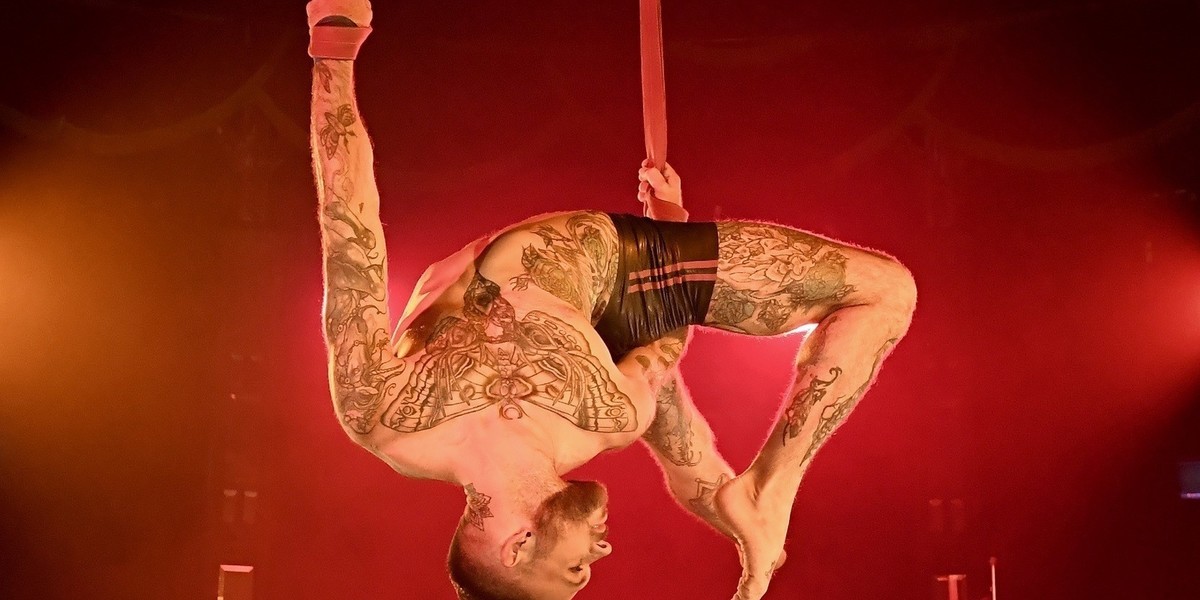 Tattooed muscly man wearing only sparkly shorts hangs upside down holding straps to suspend himself while contorting himself into a back bend