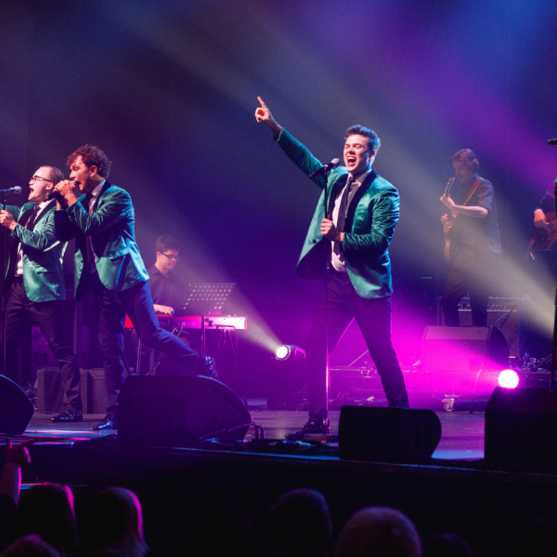 Four performers on stage in a line, singing into microphones on stands. They're dressed in green jackets, and the soloist has his hand raised and pointing out to the crowd. Behind them, are members of the band, and in the foreground are the heads of audience members.
